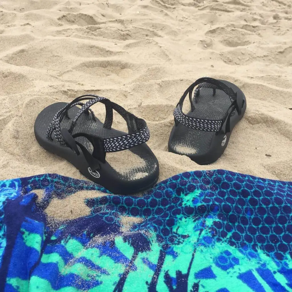 Black Chaco sandals on a sandy beach next to a blue and green patterned towel.