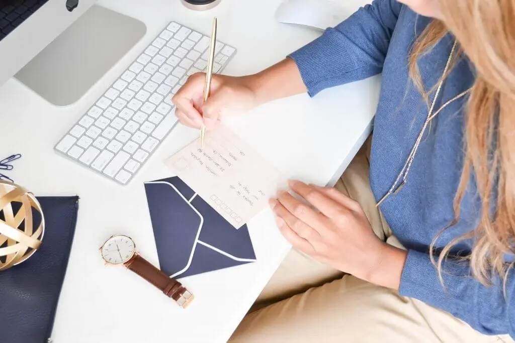 Woman writing a check at a desk with a keyboard, watch, and envelope.
