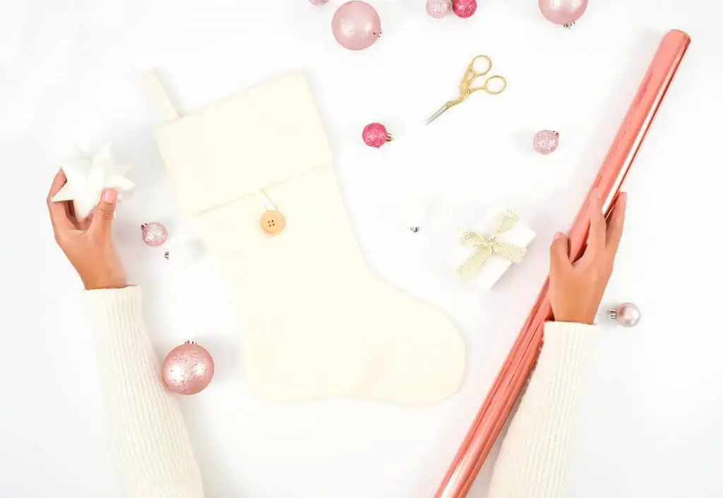 Hands wrapping a stocking with pink paper, surrounded by ornaments.