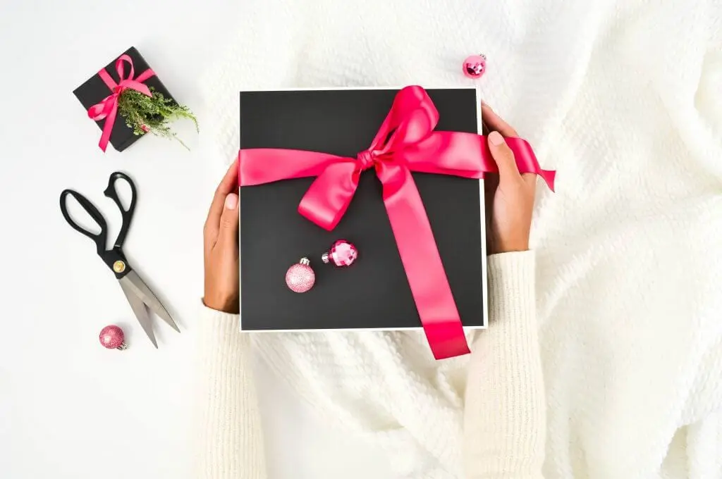 Hands holding a black gift box with a pink ribbon, surrounded by ornaments and scissors on a white background.