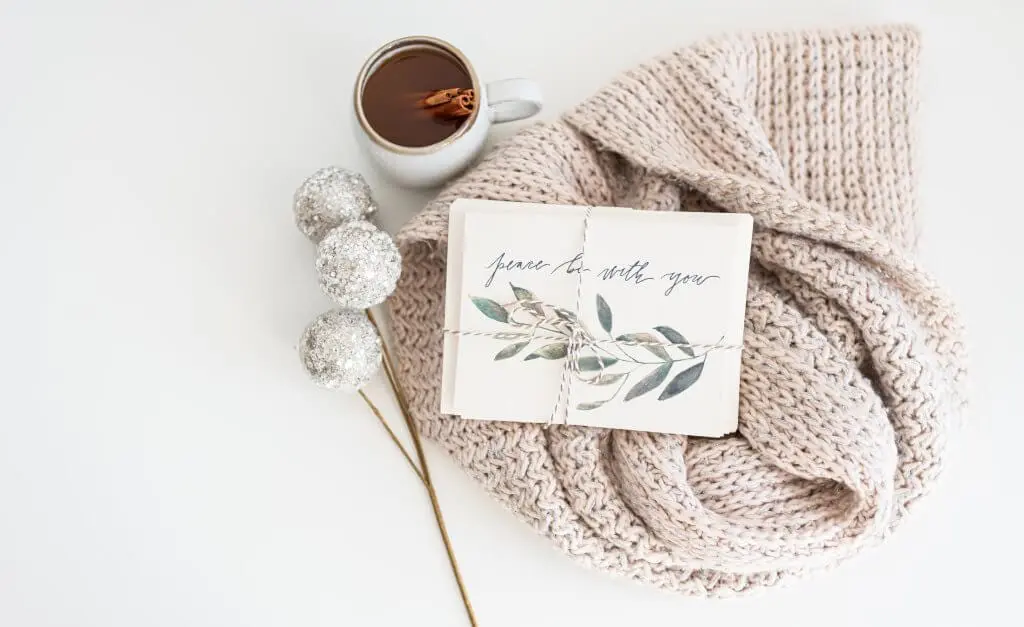 Holiday cards with "Peace be with you" text, warm drink, and cozy scarf.