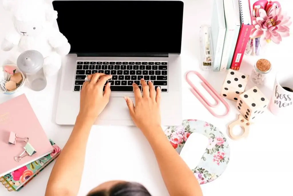 Woman typing on laptop at a bright, clean desk with "Boss Mom" mug, planner, and baby accessories.