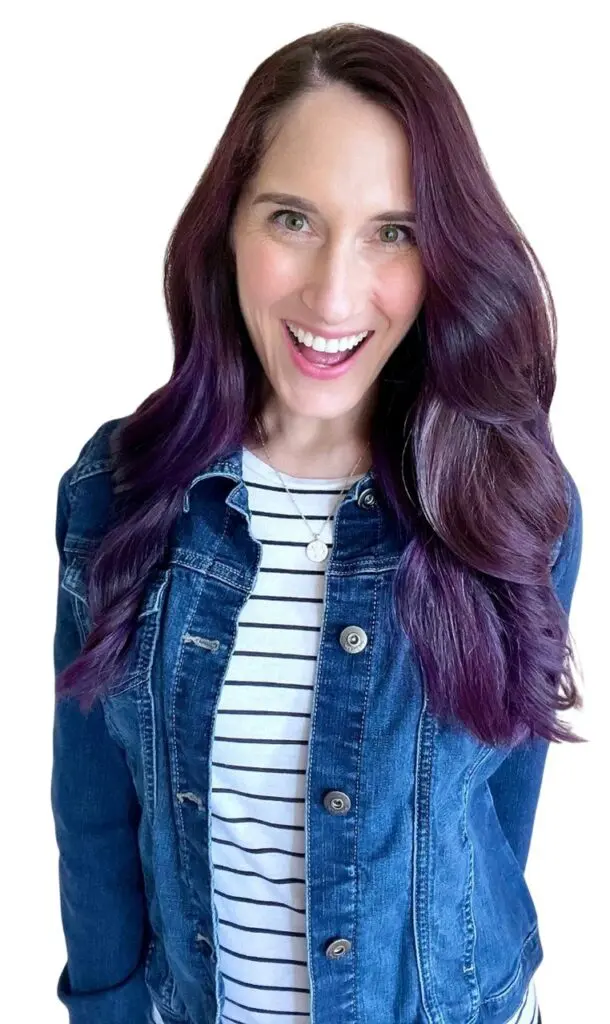 Smiling woman with purple hair wearing a denim jacket and striped shirt.