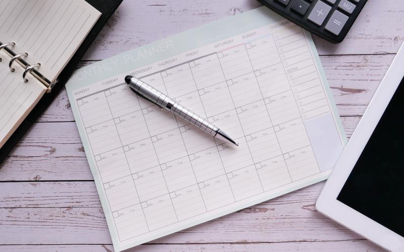 Monthly planner with pen, notepad, tablet, and calculator on a desk, planning to slow time down.