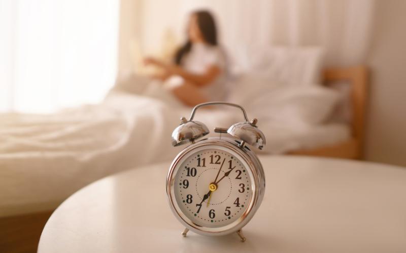 Alarm clock on a table, person sitting in bed in background. Schedule and time management concept.