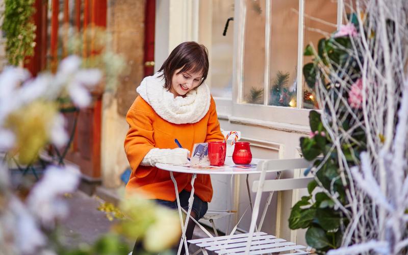 Woman writing Christmas cards at an outdoor cafe, simplifying ADHD holidays.