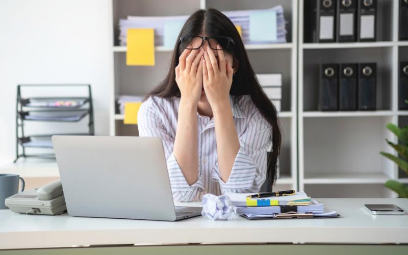 Overwhelmed woman at desk with laptop, covering face, a sign of ADHD burnout.